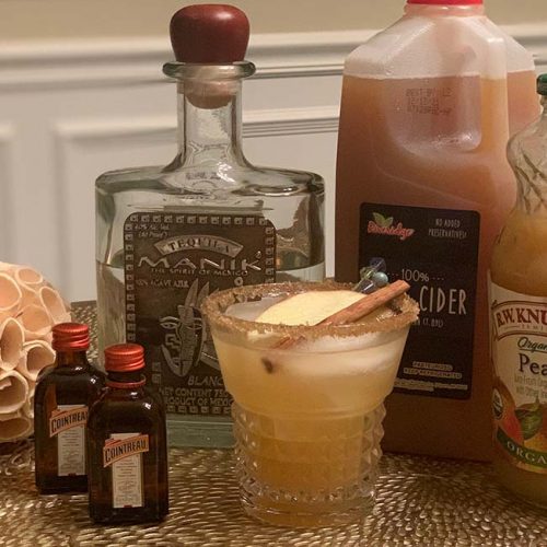 Bottles of Tequila and Cointreau Liqueur, apple cider, and pear juice, with a mixed drink in the middle, garnished with a cinnamon stick and an apple wedge.