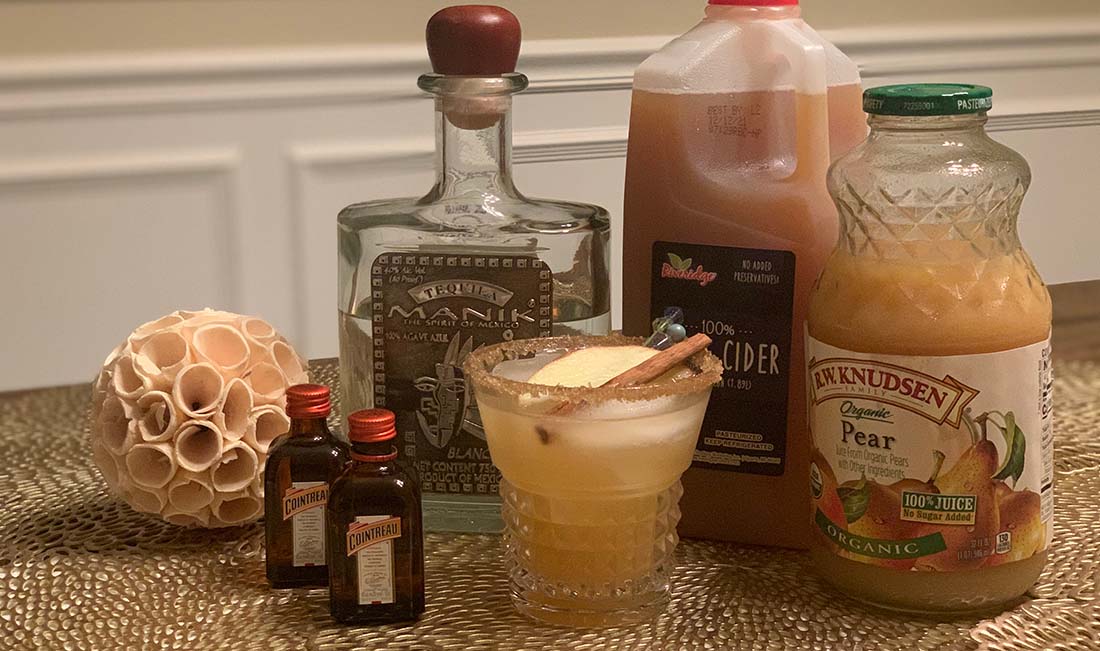 Bottles of Tequila and Cointreau Liqueur, apple cider, and pear juice, with a mixed drink in the middle, garnished with a cinnamon stick and an apple wedge.