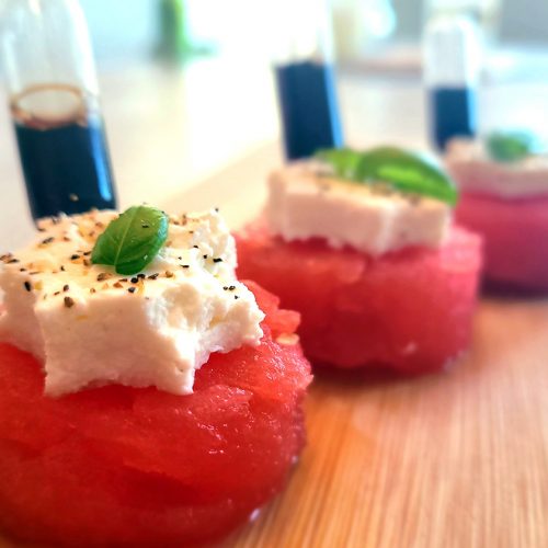 watermelon chunks with feta cheese cut in a star with a leaf of basil on top a pipette filled with balsamic vinegar is stuck in the watermelon