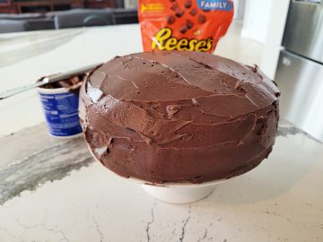 chocolate frosted cake on a white ceramic cake stand with a bag of reeses mini cups candy in the background