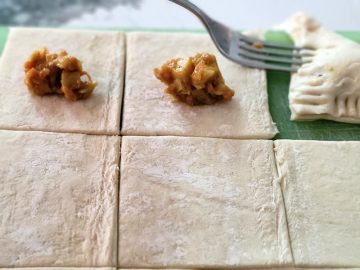 squares of uncooked puffed pastry with spoonful of curry potato, onion mixture in the middle with a fork working on closing the pastry over the mixture