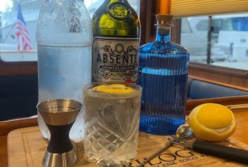 cocktail sits in front of a bottle of club soda absente and gin with a lemon a alcohol measuring jigger