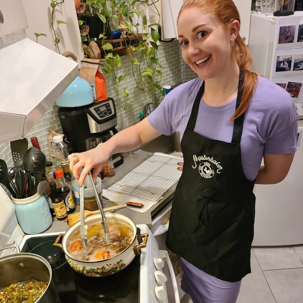 young woman with red hair in front of a stove cooking wearing a purple dress and a black apron with a white logo on it