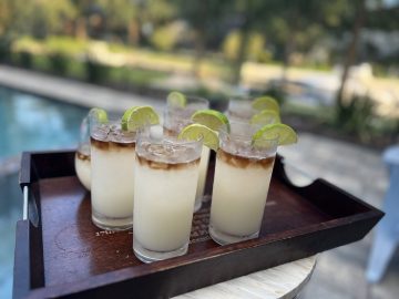 glasses near a pool with a white colored drink topped with a floater of a dark liquor and lime garnish