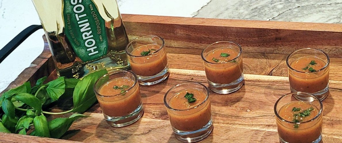 6 shot glass on a platter filled with red gazpacho with a bottle of horitos tequila behind them