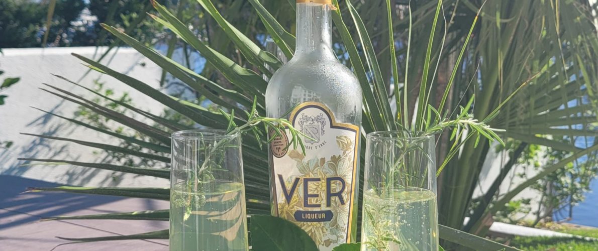 VER liqueur in front of palms. There are also two cocktails with rosemary garnish
